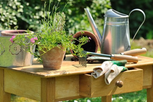 A wooden outdoor garden table is set in a lush, green garden environment with vibrant foliage in the background. The table displays several gardening tools and accessories, including a galvanized metal watering can, a pair of gardening gloves, a small garden fork, and a wooden-handled trowel. There are three plant pots on the table, one contains a small flowering plant with pink flowers, while the others appear to be empty or contain soil. The surface of the table shows natural wood grain and textures, and the overall scene suggests a well-maintained outdoor space suited for garden maintenance and landscaping tasks. This setup is indicative of a gardening activity, with the tools and pots positioned for easy access, reflecting the practical aspects of garden care in the Raynes Park area. Bright, natural sunlight illuminates the scene, emphasizing the fresh, natural tones of the plants and wooden furniture, suitable for a gardening service webpage for hedge trimming and outdoor maintenance in the local area.