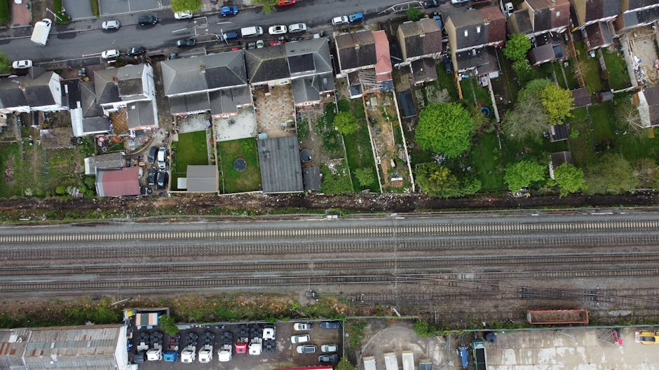 An aerial view of a residential area showing a row of back gardens alongside a railway track. The gardens feature a variety of landscaping elements, including well-maintained lawns, flower beds, and p