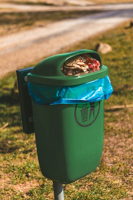 A green outdoor waste bin with a blue plastic bag lining and a transparent lid, filled with assorted garden waste such as paper, plastic, and organic debris. The bin is situated on a grassy area near 
